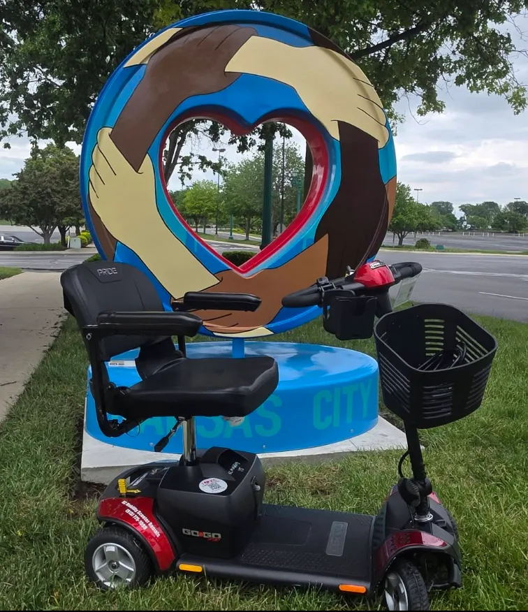 A red Pride Go-Go mobility scooter parked in front of the Kansas City hands-around-heart sculpture, with the words KANSAS CITY visible on the base.