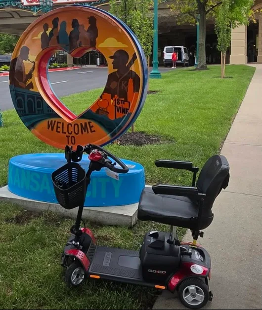 A red Pride Go-Go mobility scooter parked beside the 'Welcome to Kansas City' heart sculpture at 18th & Vine, which depicts silhouettes of jazz musicians and the words 18TH VINE.