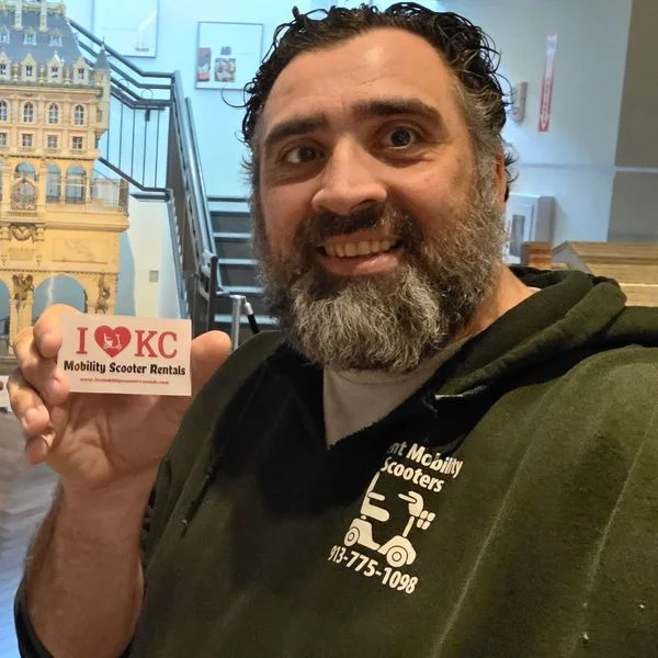Jeff holding a KC Mobility Scooter Rentals card in a Kansas City museum gallery of miniature buildings.