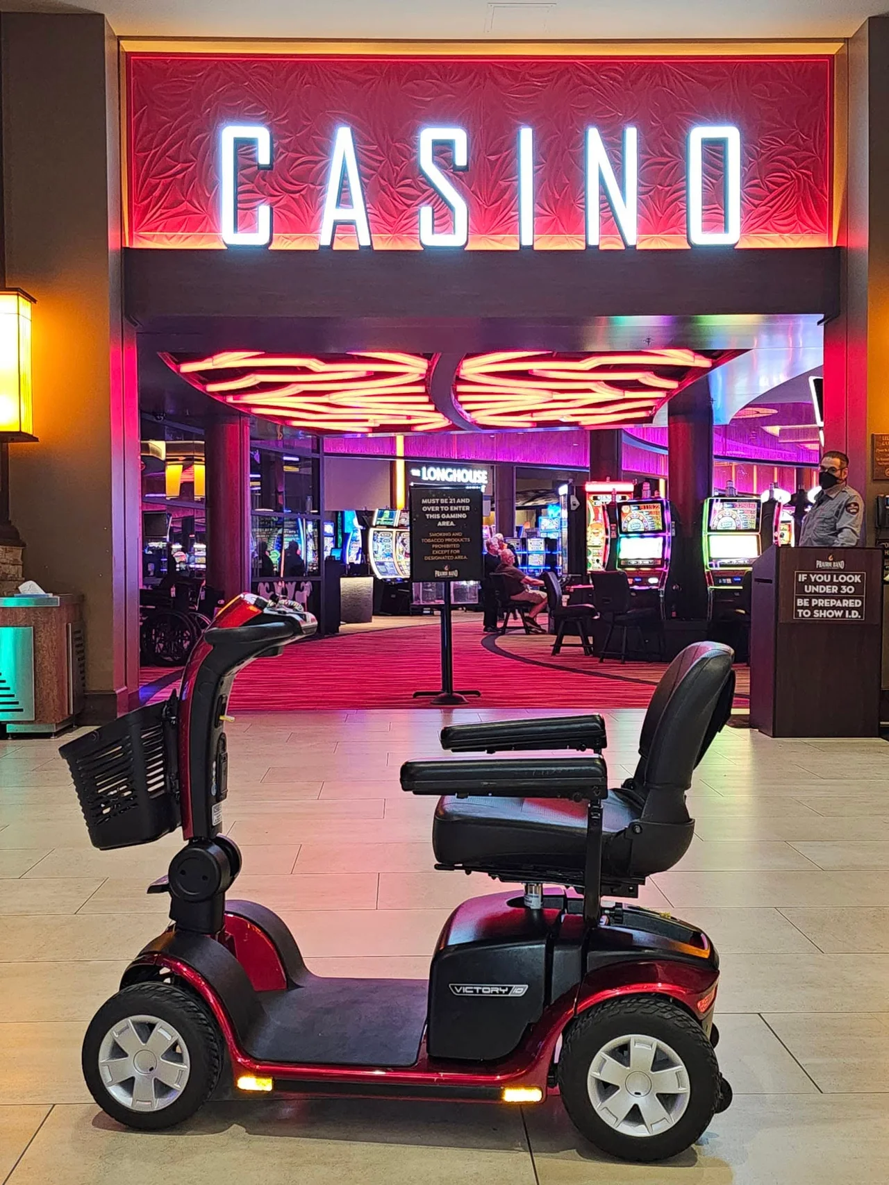 Pride Victory 10 four-wheel mobility scooter in Candy Apple Red staged at the gaming-floor entrance of Prairie Band Casino, illuminated CASINO sign overhead, awaiting customer pickup.