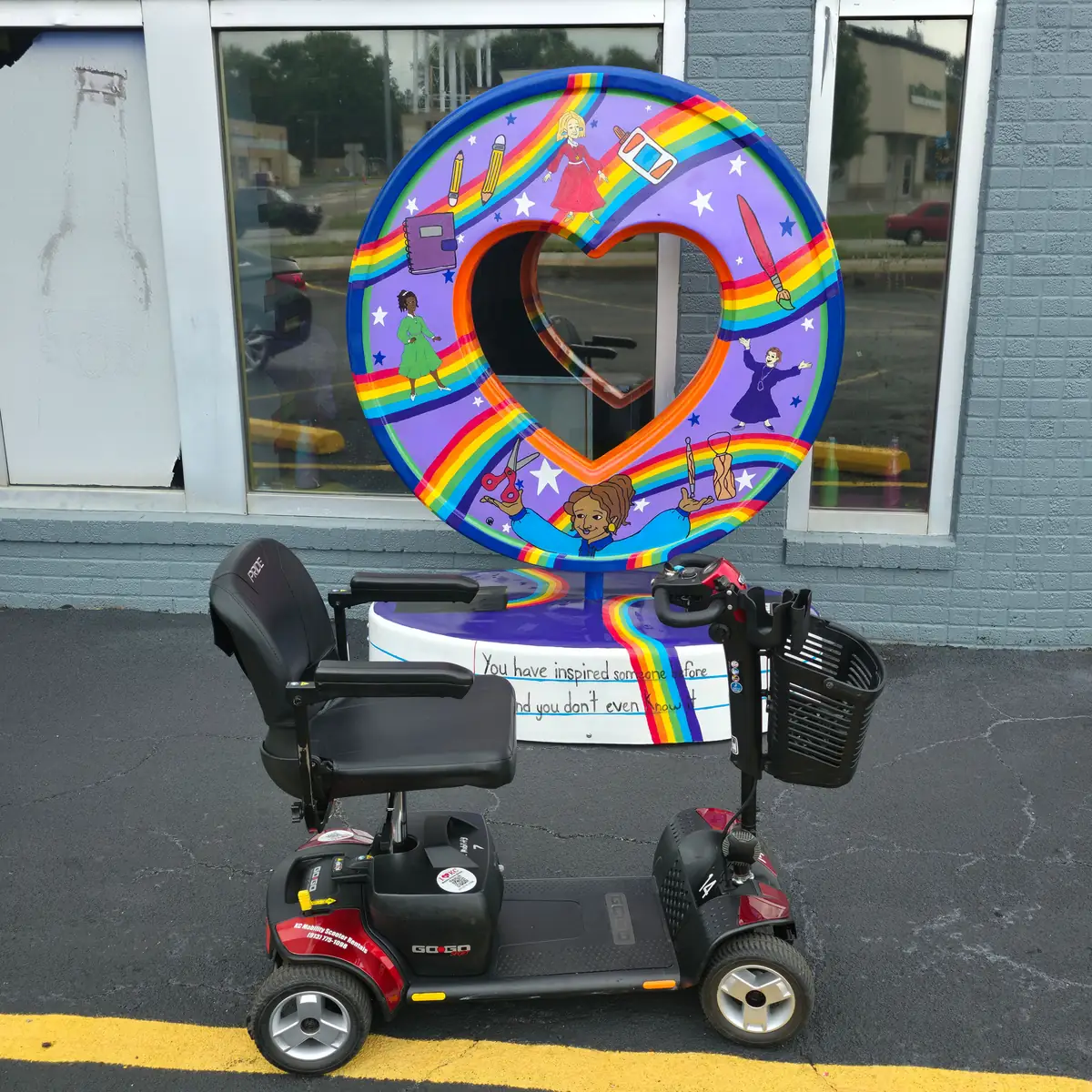 Red Pride Go-Go Traveller 4-wheel mobility scooter in front of a rainbow-painted heart sculpture in the Kansas City area