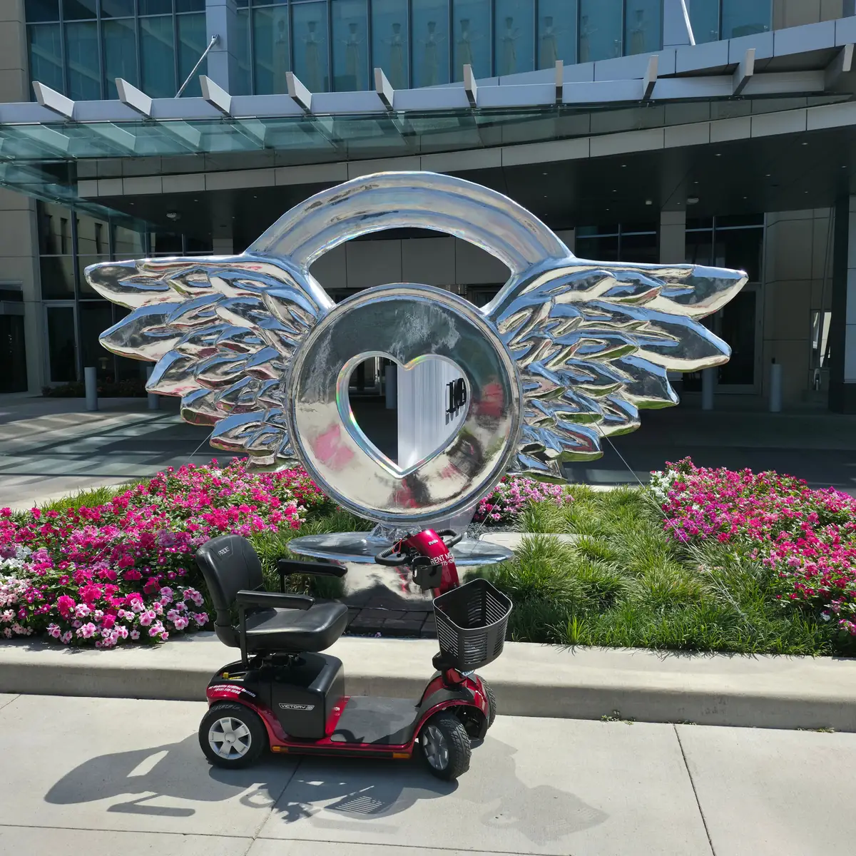 Red Pride Victory 10 4-wheel mobility scooter in front of a silver winged heart sculpture and flower bed in downtown Kansas City