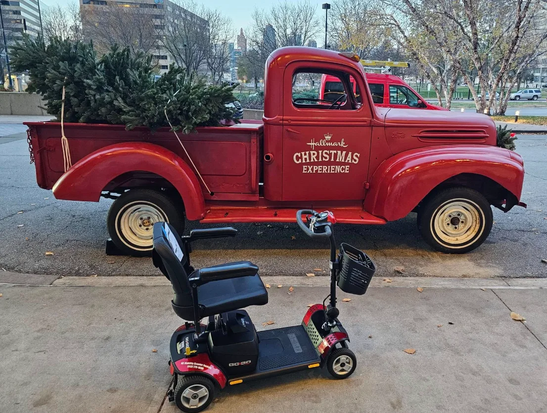 Pride Go-Go mobility scooter beside the Hallmark Christmas Experience vintage red truck with a tree in the bed, downtown Kansas City skyline in the background