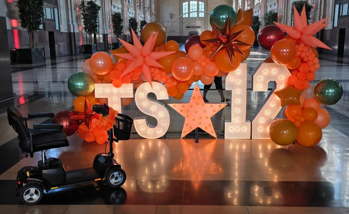 Pride mobility scooter staged in front of an illuminated marquee letter installation and orange balloon arch inside a Kansas City venue lobby