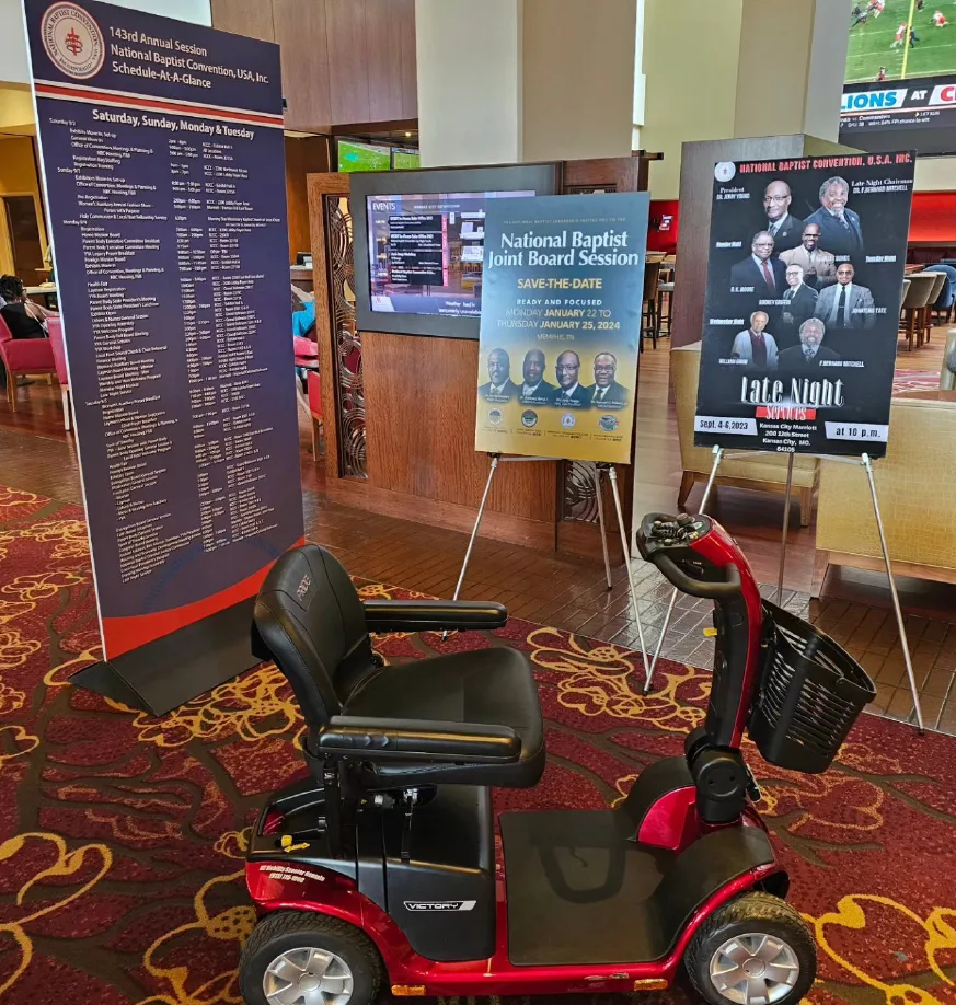 Pride Victory 10 mobility scooter staged in a Kansas City convention hotel lobby beside a multi-day session schedule board, a save-the-date easel, and event signage