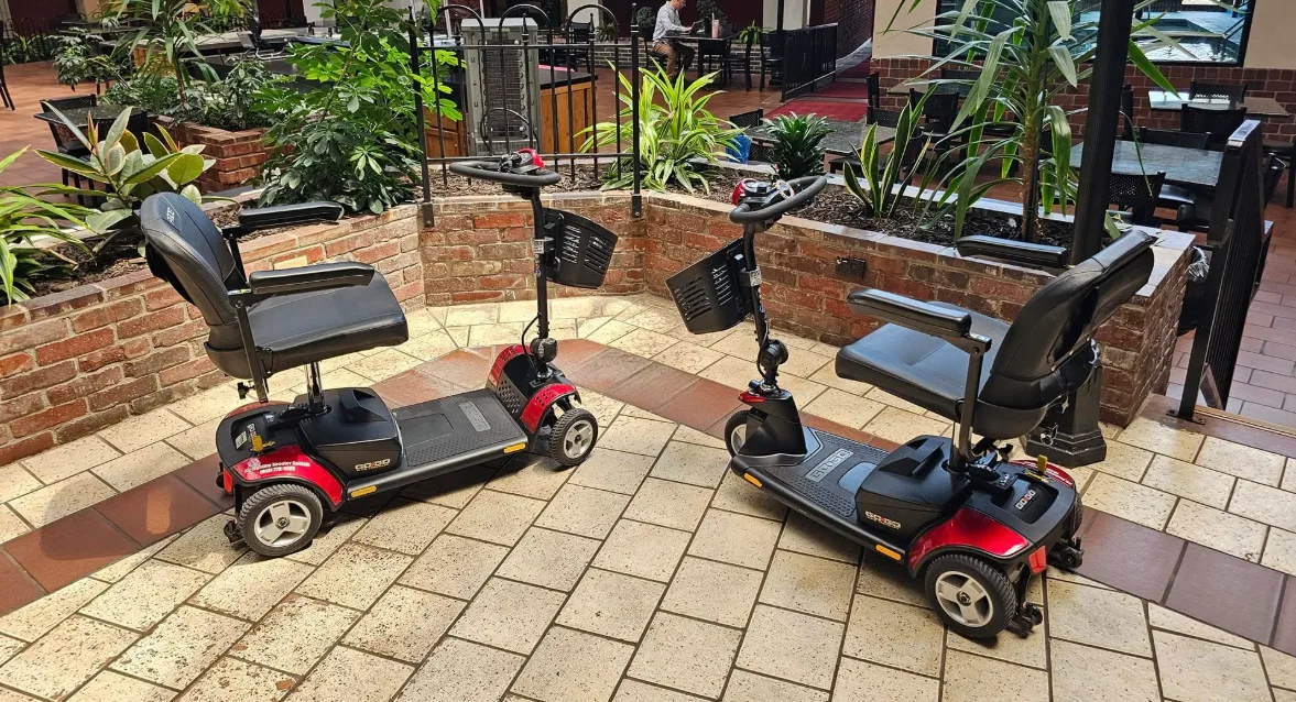 Two Pride Go-Go mobility scooters staged side-by-side in a hotel atrium courtyard, ready for bell-stand handoff to incoming guests