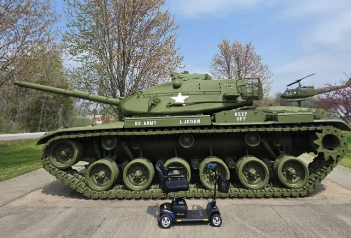 Two KC Mobility Scooter Rentals mobility scooters parked in front of a vintage U.S. Army tank memorial display