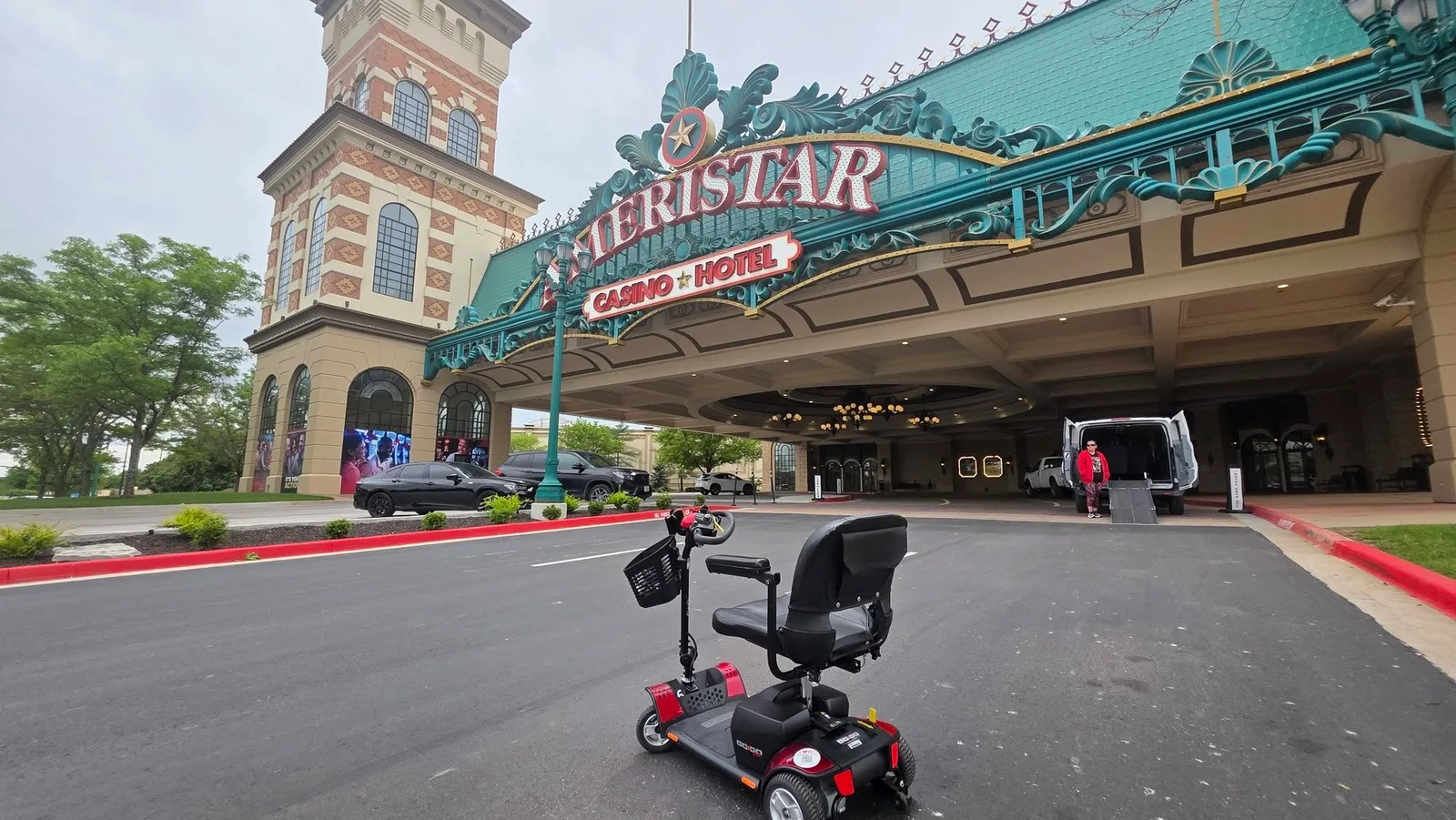 Pride Go-Go red mobility scooter staged on the porte-cochère drive at Ameristar Casino Hotel Kansas City, with the casino's signature teal-and-gold marquee overhead and a wheelchair-accessible ramp van unloading at the valet curb in the background.
